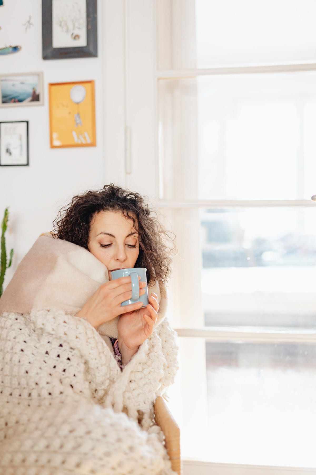 A woman wrapped in a cozy knit blanket sipping from a mug in a bright living room