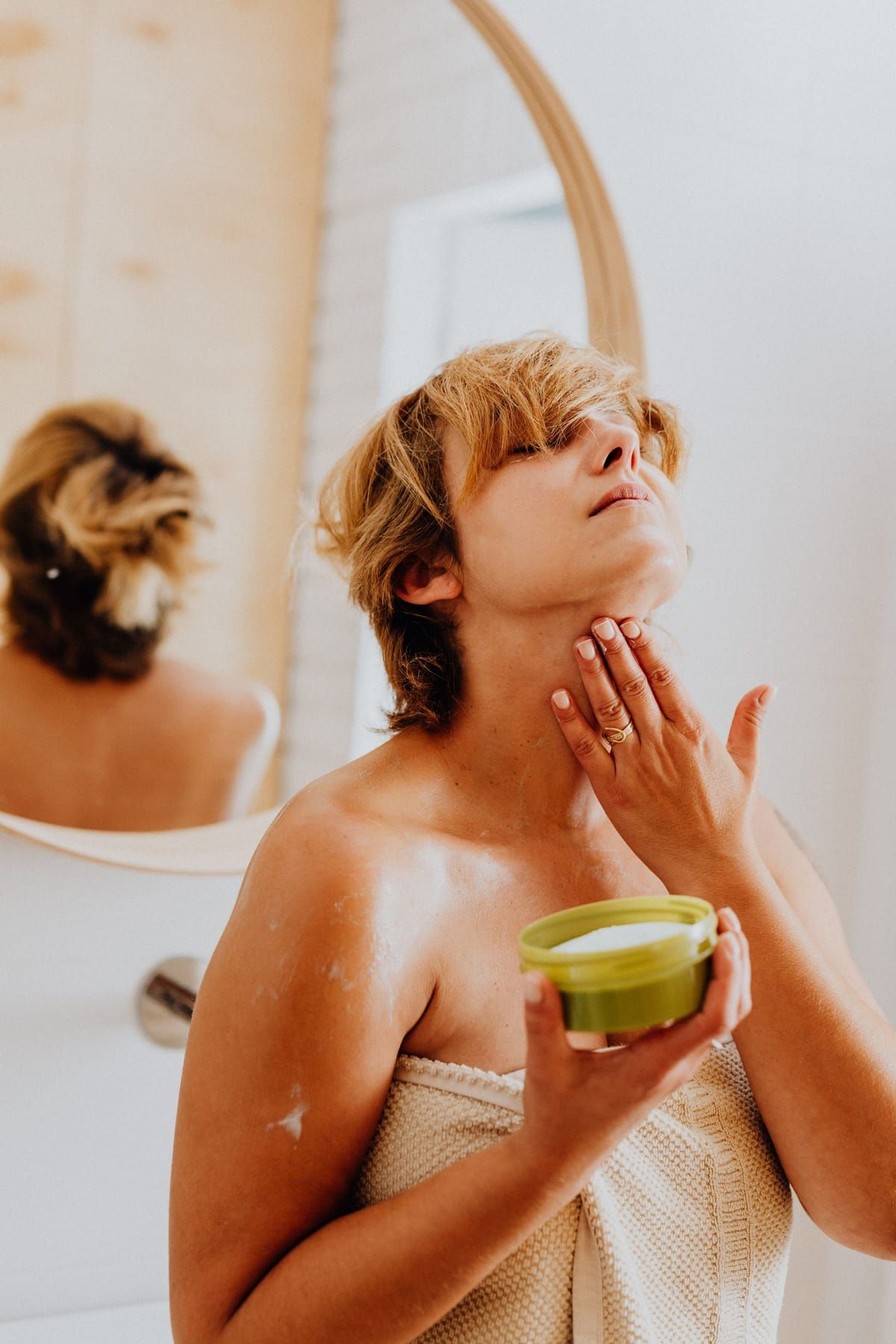 A woman wrapped in a towel smoothing cream from a small jar onto her neck in front of a bathroom mirror
