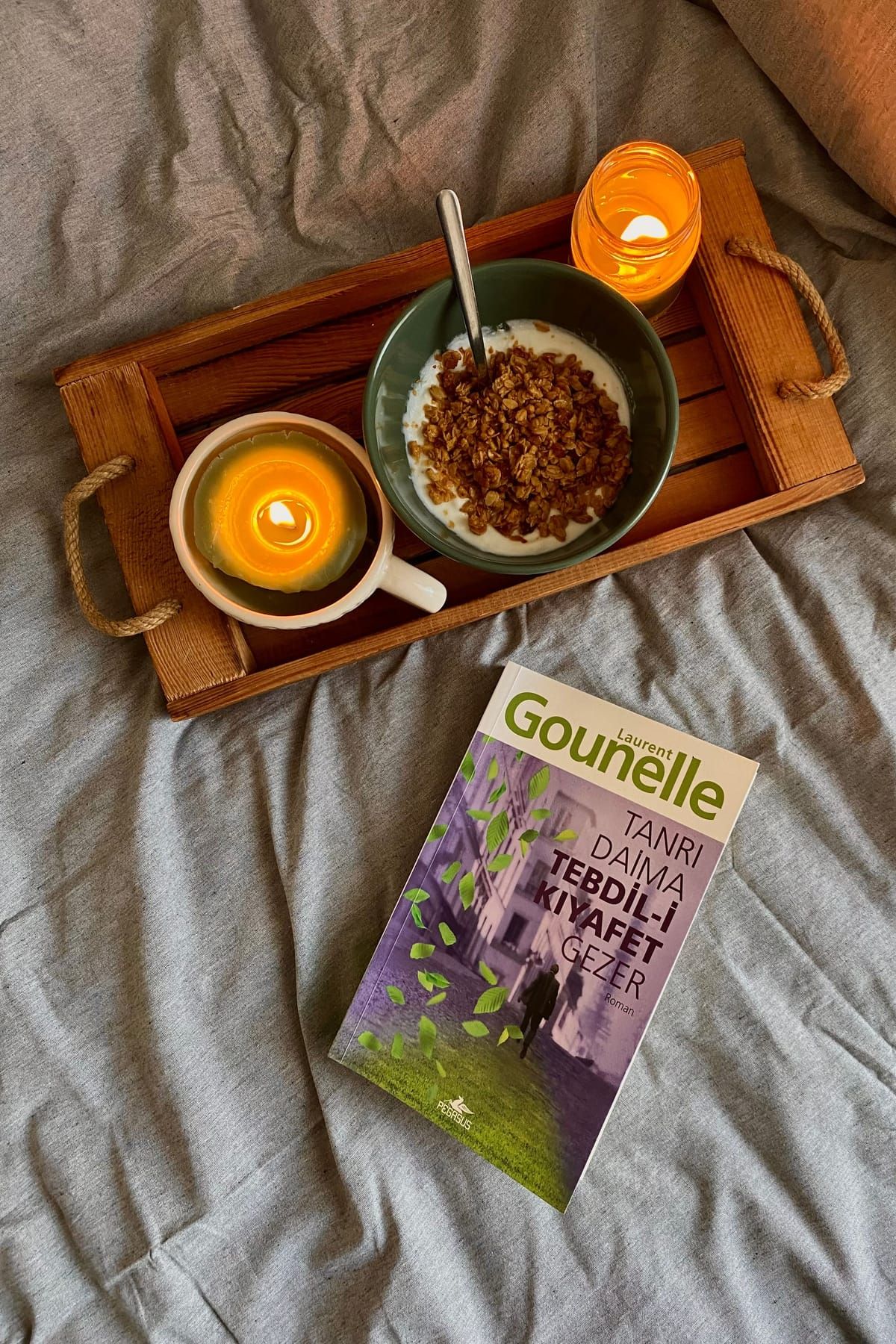 A wooden breakfast tray with a lit candle, a mug and a bowl of granola on a bed with soft gray sheets, beside a book