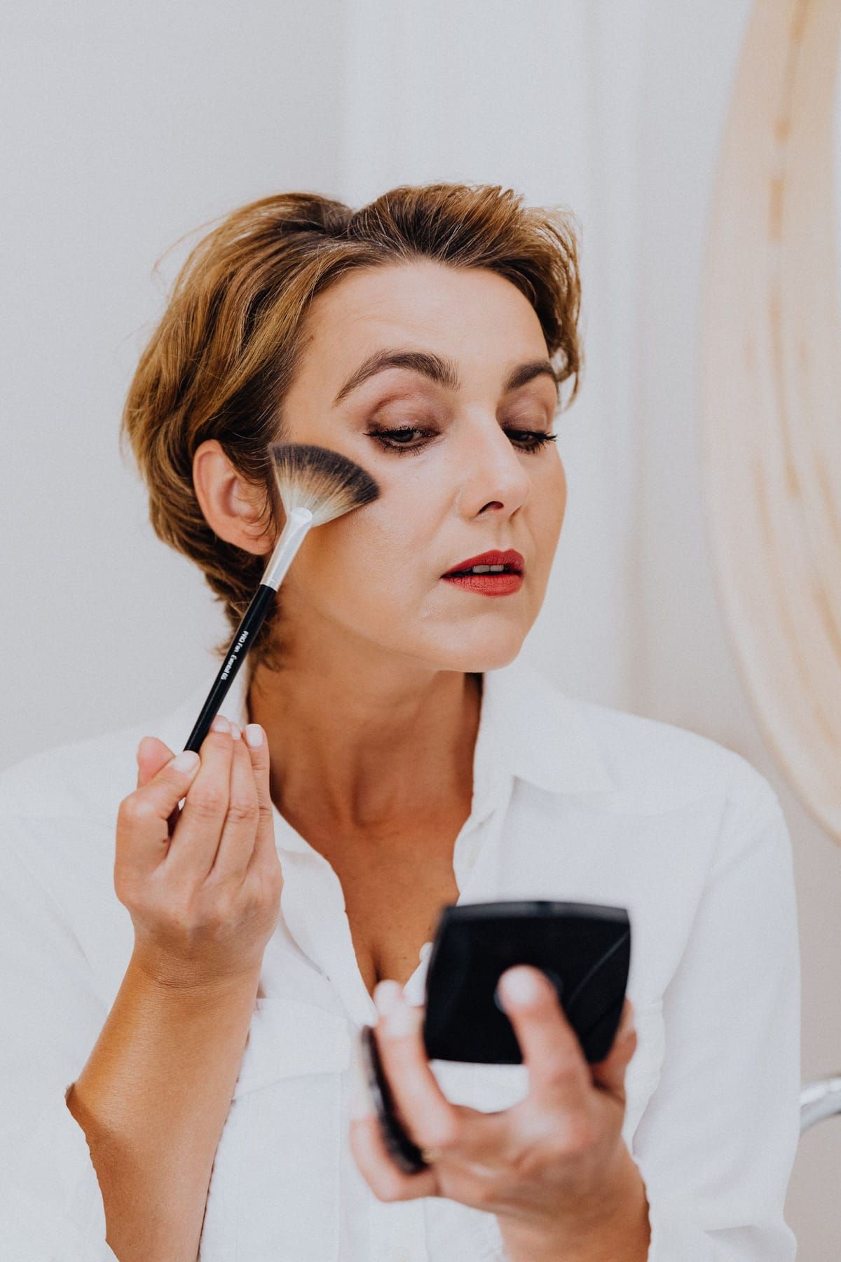 A woman sweeping blush onto her cheek with a brush while holding a compact mirror