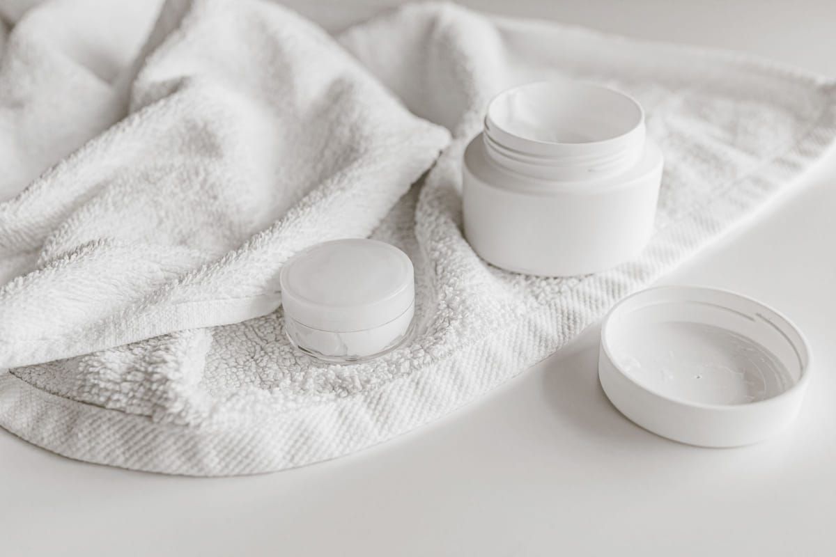 White skincare jars with their lids off, resting on a soft white towel